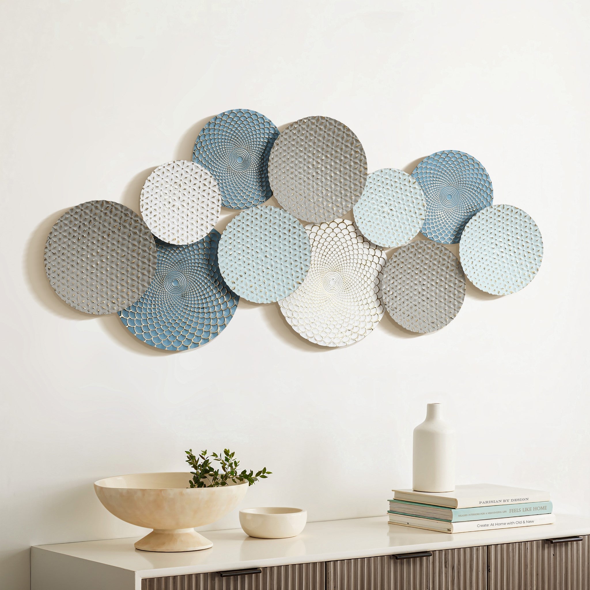 A vintage metal wall decoration, this abstract wall art showcases a series of overlapping circular metal discs in hues of blue, gray, and white with intricate patterns, elegantly mounted above a cabinet adorned with decorative items.