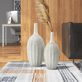 Two LuxenHome modern geometric pattern resin trumpet vases, 23.5 h, stand on a rug in the living room. One holds tall pampas grass. The wooden wall background includes a desk with a computer and part of a sofa.