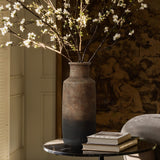 A LuxenHome Brown Ombre Terracotta Tall Vase, 14.8 h, with blossoming branches sits on a round table. Nearby, a small stack of books rests in soft light against the backdrop of a muted tapestry, creating an elegant and serene minimalist decor.