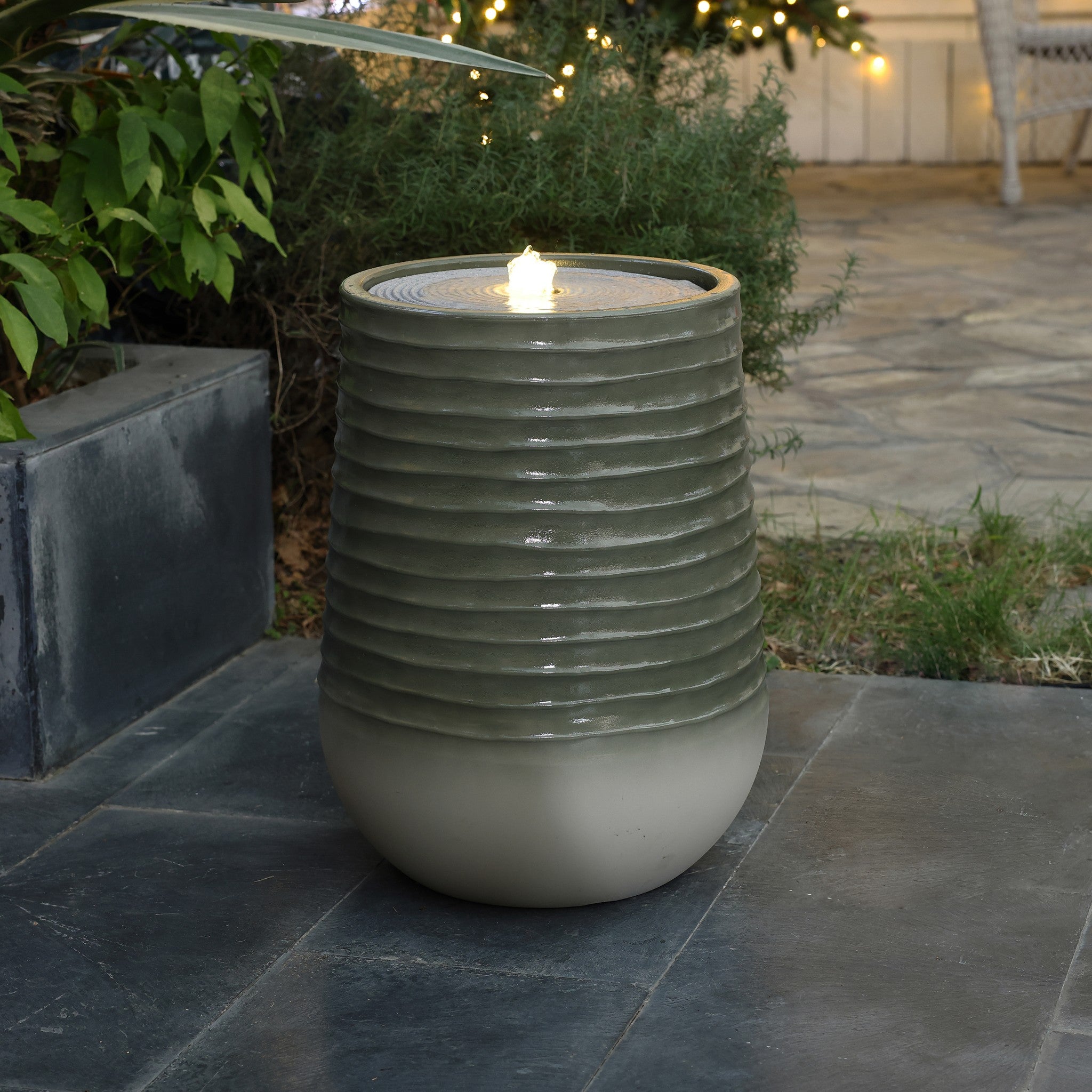 A ceramic outdoor fountain with a fluted design graces the patio, its central spout gently flowing water. Crafted from high-density resin and accented by LED lights, it shines softly among potted plants and the paved stone flooring.