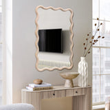 A wavy-framed decorative wall mirror reflects a wall-mounted TV and radiator. Below, a console table with a natural wooden frame holds a vase of dried flowers, a bowl, and a stack of books titled Best Furniture.