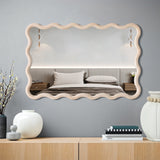 A wavy-edged, decorative wall mirror reflects a bed with a neutral-toned headboard and pillows. On the dresser with a pine wood finish below, there are decorative vases, a stack of books, and a framed photo.