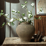 The LuxenHome rustic brown terracotta cauldron vase, 14.6 h, with two handles holds branches of white flowers and green leaves on a wooden table beside stacked logs. A framed artwork and textured wall in the background complete the elegant scene in natural daylight.