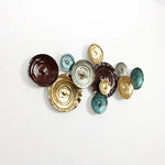 Wall art featuring an abstract design of textured, circular ceramic pieces in brown, gold, and teal hues, mounted against a white background.
