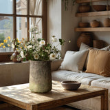 A rustic living room features a large cement cylinder vase with white flowers on a wooden coffee table, a beige sofa with cushions, and wooden shelves showcasing pottery with a ginger root brown finish, all bathed in natural window light.