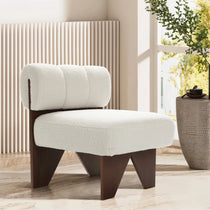The LuxenHome Lorenzo armless accent chair, with its white boucle upholstery and dark wooden frame, exudes mid-century charm in a sunlit modern room with wooden flooring. Beside it sits a basket holding a cup and saucer, while large windows reveal lush green foliage.