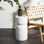 A modern outdoor scene features a woven wooden chair beside the sleek Speckled White Pillar Outdoor Side Table. On the white cylindrical table, made from durable MGO material, sit a bottle of Jack Daniels whiskey and a glass. Greenery gently casts shadows on the nearby wall.