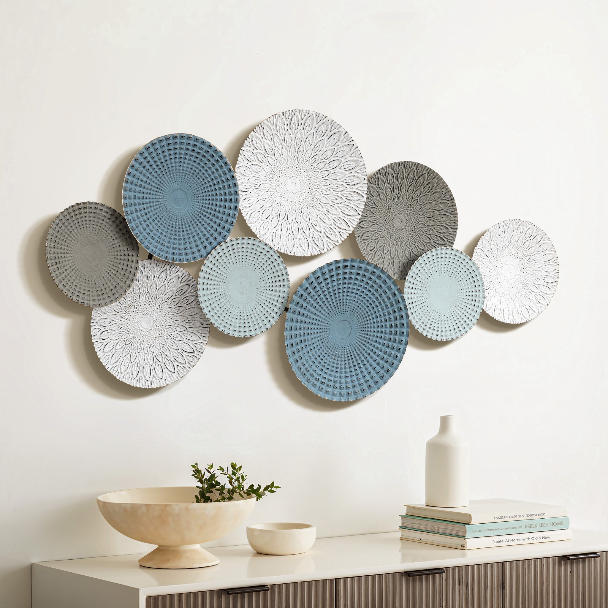 A collection of decorative plates with intricate patterns is mounted on a wall above a wooden sideboard, adding an artistic touch to your home decor. Nearby, an abstract metal wall decoration complements the ceramic bowl with a small plant and a stack of books on the sideboard.