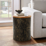 A round black and gold table with a hammered iron base stands beside a beige sofa, holding a small bowl and a glass of ice water, in a sunlit room with a parquet floor.