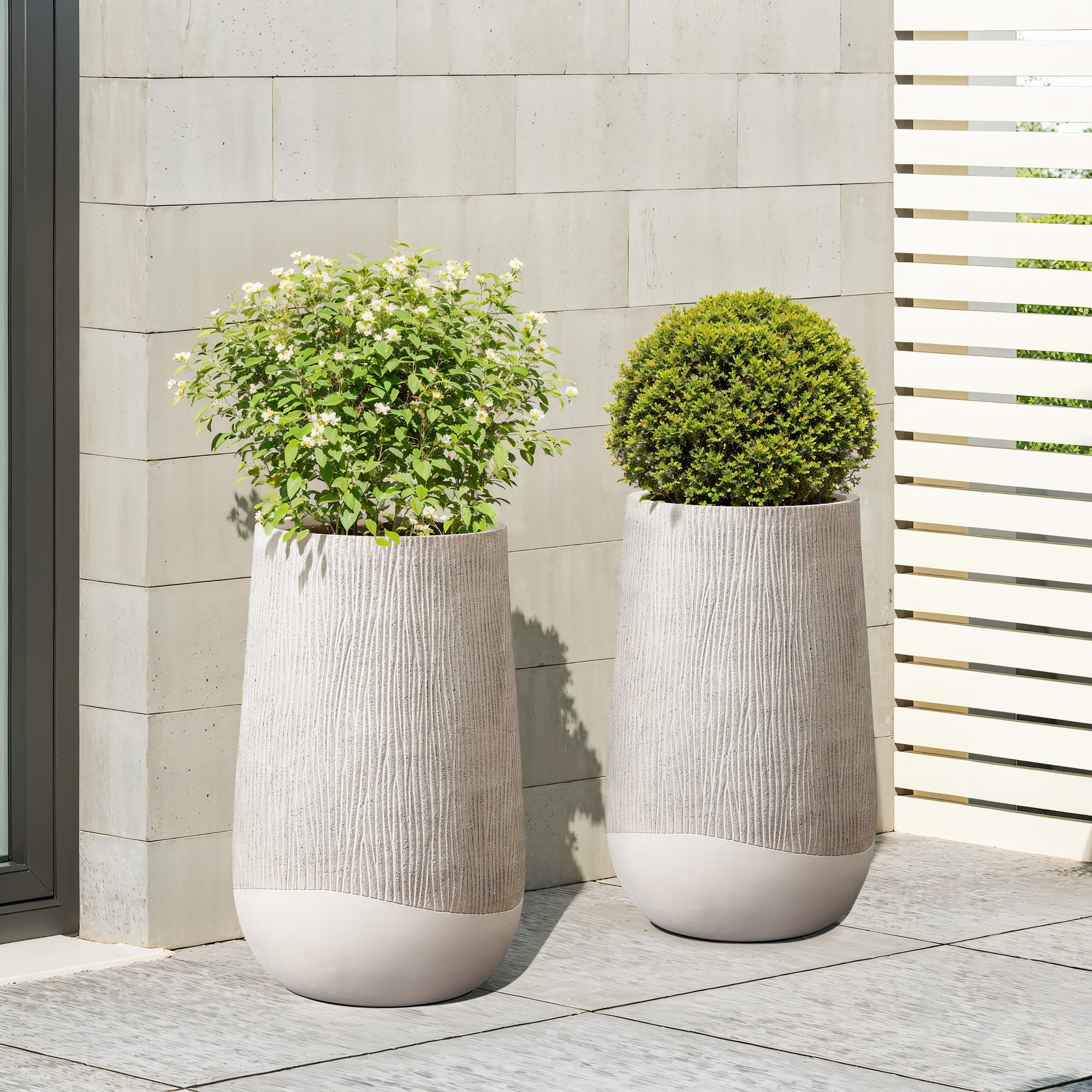 Two tall, textured planters enhance the patios modern minimalist aesthetic. The left planter, with a natural wood pattern, cradles a bushy plant with small leaves, while the right round tapered planter showcases a neatly trimmed shrub.