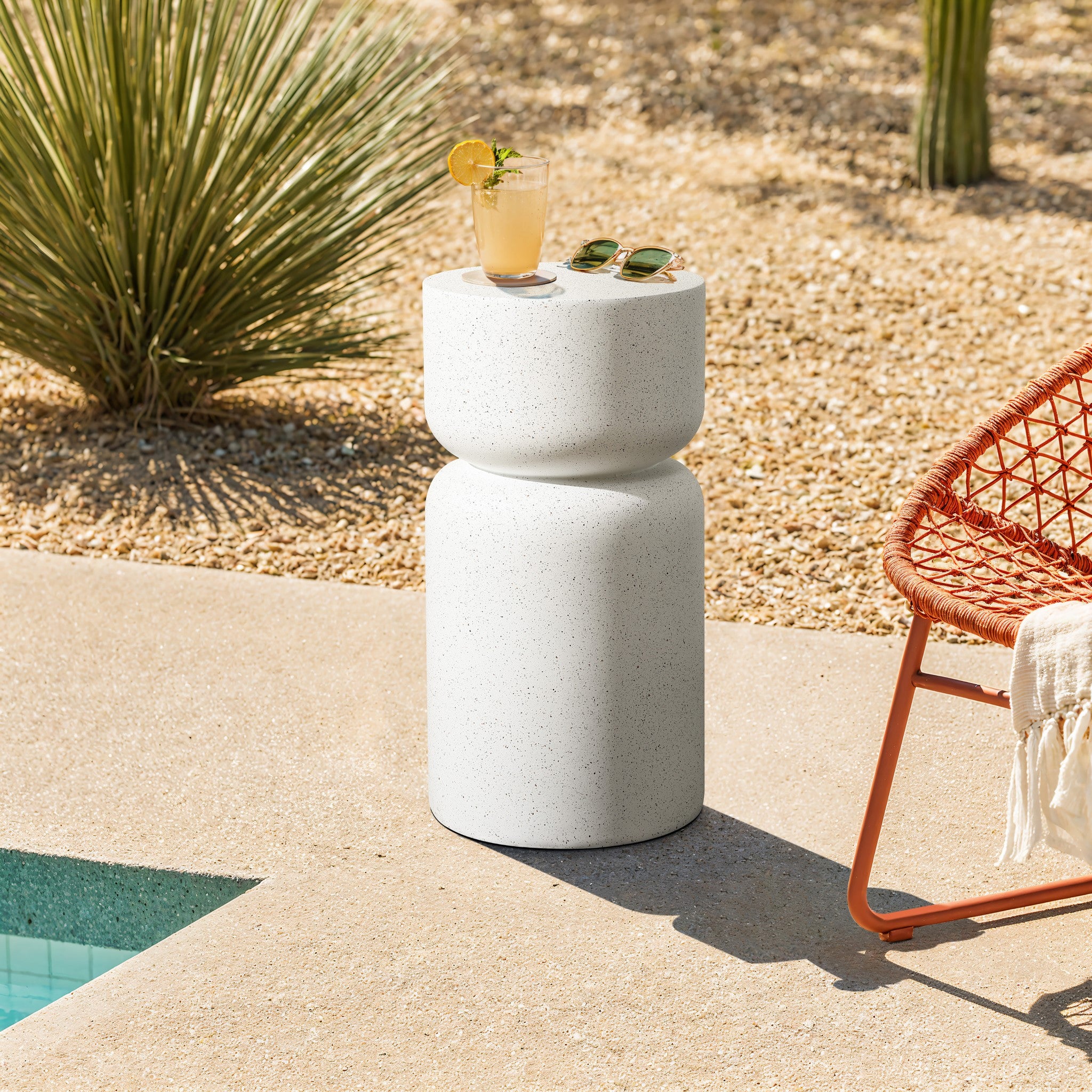 A stylish outdoor scene showcases a speckled white pillar outdoor side table by the pool, crafted from durable MGO material. It holds a glass of lemonade and sunglasses. A red woven chair partially covered by a white blanket sits nearby, surrounded by desert plants.