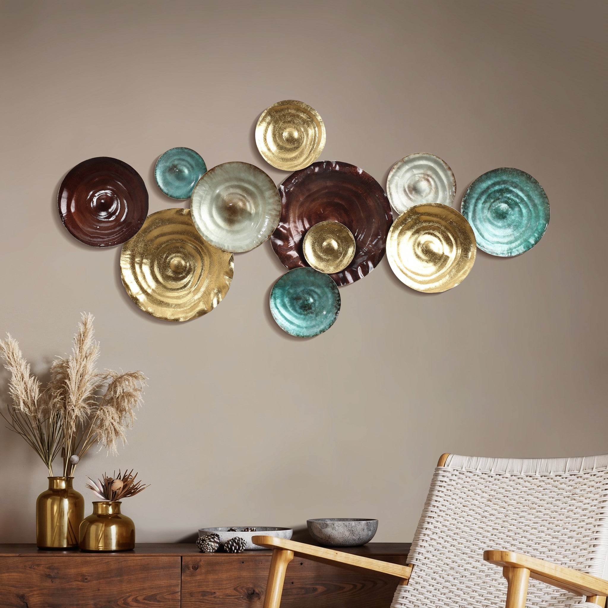 Metal wall art featuring metallic circular plates in gold, bronze, brown, and teal adorns a beige wall above a wooden cabinet. Pampas grass in vases and a wicker chair complement the abstract design.