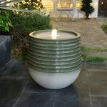 A round, modern fountain with a ribbed ceramic design features LED lights glowing softly from its center. It sits outdoors on a stone patio surrounded by plants, with a house visible in the background.