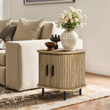 A cozy living room corner features a beige sofa adorned with patterned cushions. LuxenHomes Aria oak finish nightstand with a slatted sliding door holds a vase of white roses and decorative bowls, all set on a patterned rug. Natural light streams in from nearby sliding doors.