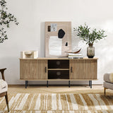 A modern living room showcases the LuxenHome Aria oak finish 55.1-Inch wide TV stand, featuring sliding doors and black legs for enclosed storage. Minimalist art and a vase with greenery adorn the top, enhancing the mid-century modern style with a patterned rug, cozy armchair, and corner plant.