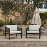 Two cushioned outdoor armchairs on a patio with a stone wall and trees in the background. Featuring PE rattan craftsmanship, these chairs promise durability. A closed patio umbrella stands nearby.