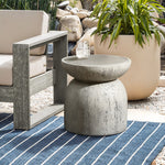 A modern outdoor setup with a rustic-style Rustic Off White and Gray Outdoor Side Table, 15.75 h, hosting a glass of water beside a light gray cushioned chair. A potted plant sits behind them on a blue and white striped rug over the stone floor.