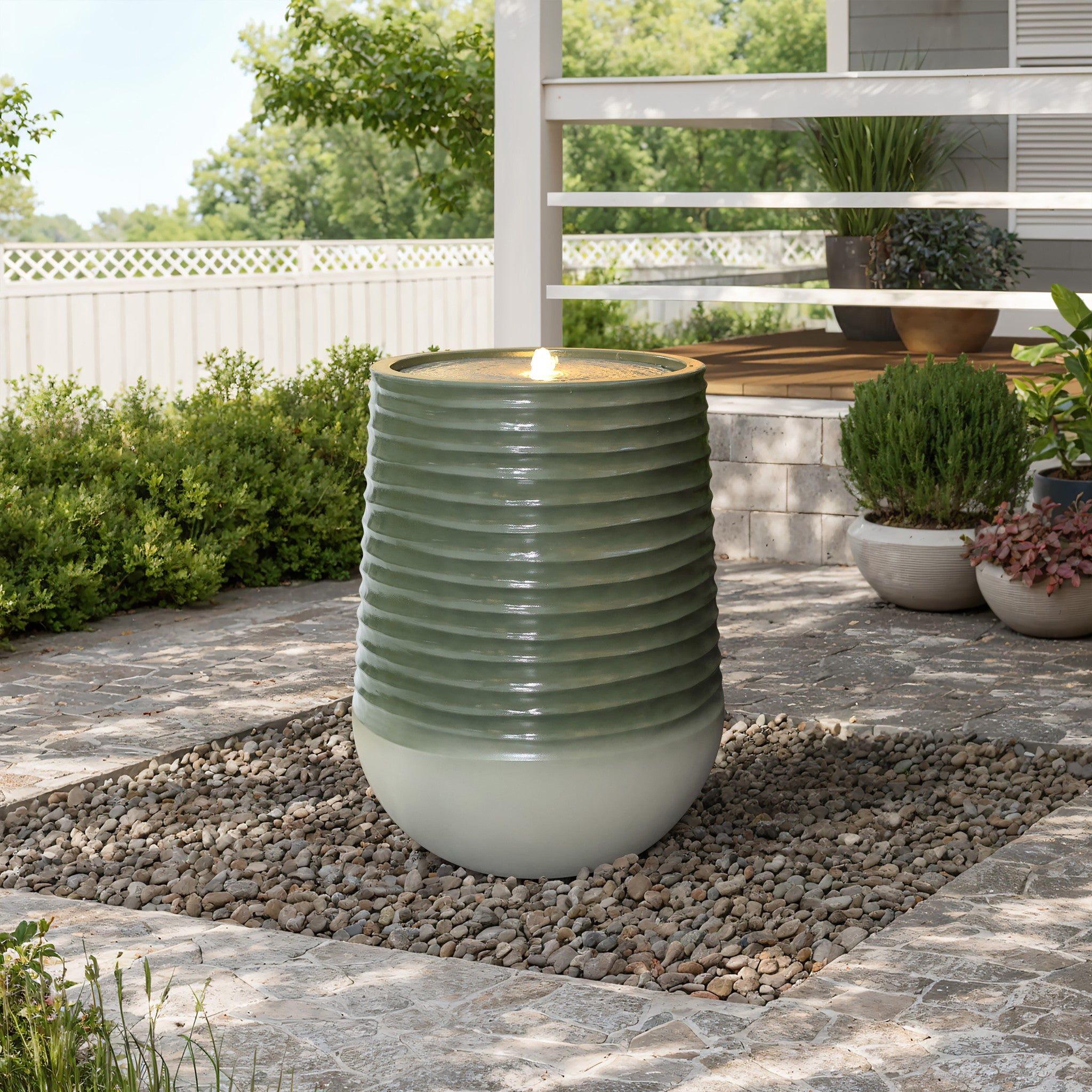 A modern fountain with a ribbed, green ceramic finish graces the gravel bed, surrounded by a stone patio and lush plants. Subtle LED water features illuminate the scene as a rustic bench offers tranquil views in the background.