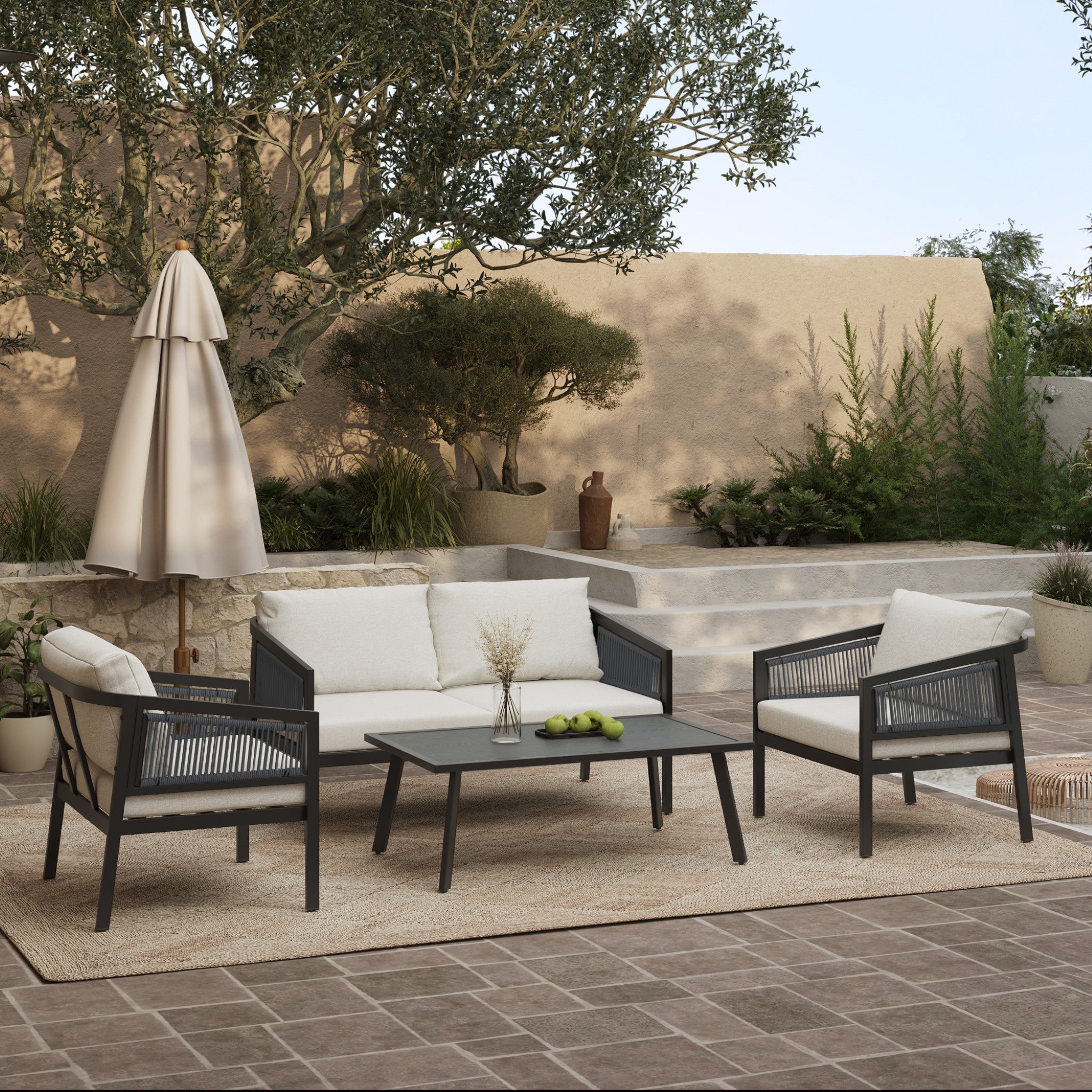 Outdoor patio set featuring an outdoor loveseat and two chairs with a sleek black steel frame. The coffee table, adorned with green apples, sits on a woven rug. In the background, potted plants, an umbrella, and stairs against a beige wall complement the PE rattan furniture ensemble.