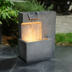 A modern, gray concrete water fountain with two tiers and integrated LED lighting, placed on a stone surface surrounded by greenery. This outdoor relaxation fountain perfectly complements the serene setting.