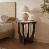 A modern bedside setup displays the LuxenHome Sunflower wood end table with curved legs. Atop is a beige ceramic lamp with a conical shade. The background features a bed with white linens and a plant on the right, showcasing seamless modern design.