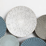 Decorative plates with intricate patterns add an artistic touch to your home decor; one central plate features a white floral design, surrounded by others in blue, grey, and green tones with textured patterns. Its like an abstract metal wall decoration for your walls.