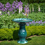 A bird with outstretched wings lands on a ceramic turquoise birdbath, nestled amidst green shrubs and purple flowers, enhancing the tranquility of the outdoor space.