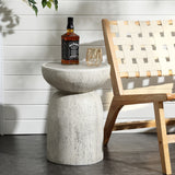 A patio scene includes a woven chair next to a LuxenHome Rustic off White and Gray Outdoor Side Table. On the table, theres a bottle of Jack Daniels whiskey and an empty glass. The setting features a white shiplap wall with green leaves hanging down.