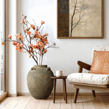 In a cozy room, a wooden chair with an orange-patterned pillow sits beside a small table with a white mug. Elegantly placed is the LuxenHome Rustic brown terracotta cauldron vase at 15.4 inches tall. A framed tree branch print decorates the white wall, enhancing the natural setting.