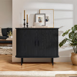 The Aria 2-door tambour accent cabinet in a black finish features vertical grooves and enhances a mid-century modern living room with a herringbone floor. Decorative candle holders, framed art on top, and a nearby plant on the right, alongside a couch and rug on the left, complete this stylish storage solution.