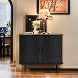 The LuxenHome Aria accent cabinet in black, featuring a mid-century modern curved design and tambour doors, stands against a cream wall. It holds a floral-patterned lamp, framed picture, books, and flowers on top. An archway in the background leads to a room with a chair and blinds.