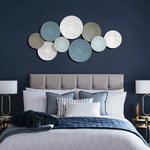 The bedroom boasts a tufted headboard, decorative pillows, and a blue quilt. Two lamps on nightstands flank the bed while the wall features an artistic touch with abstract metal wall decorations, adding color and texture to the space.
