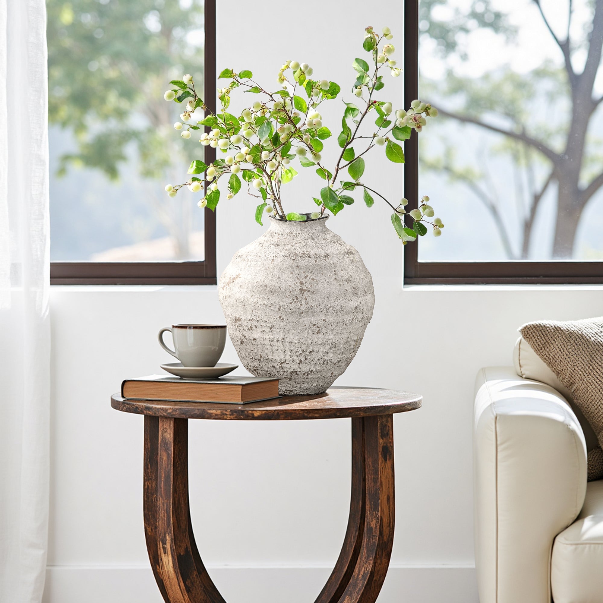 Textured cement vase holding green branches, a stacked book, set beside a white sofa in front of large windows.