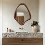 A bathroom featuring a textured stone countertop and wooden cabinet embodies Scandinavian charm with its unique wooden-framed wall mirror, gold faucets, and a minimalist design accentuated by a vase with flowers.