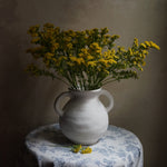 yellow florals in a white ceramic vase with 2 ears on top of a table/stool covered by printed blanket