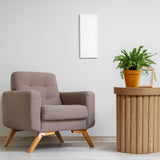 A beige armchair sits beside a round wooden end table with an oak finish, featuring a potted fern and a framed photo against a light gray wall.