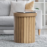 A round wooden end table with an oak finish and fluted drum design stands on a gray carpet beside a light gray sofa. A laptop and stacked books are on the table.