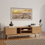 A modern living room showcases the LuxenHome Eric oak finish 70 TV stand adorned with decorative items, alongside a framed aerial cityscape at sunset. A patterned rug sits on the wooden floor, with a partial view of a plant to the right.