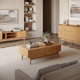 A modern living room features light wood furniture with an Eric oak finish coffee table by LuxenHome, offering storage. A gray sofa faces a wooden cabinet, abstract paintings adorn the walls, and a woven basket with flowers sits on the mid-century modern coffee table.