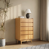 The LuxenHome Eric oak finish 5-drawer chest stands against a beige wall, illuminated in a bright room. A lamp and two books rest on top, with a tall potted plant adding greenery to the left. Curtains filter light, accentuating the Mid-Century Modern design through shadows on the wall and floor.