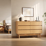 A minimalist bedroom showcases the LuxenHome Eric oak finish 6-drawer dresser, adorned with a white lamp, decor items, and framed artwork above. Sunlight casts soft shadows on the wall, with a glimpse of a bed visible in the adjoining room.