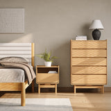 A minimalist bedroom highlights a LuxenHome Eric oak finish nightstand, beautifully showcasing a potted plant. Adjacent is a tall dresser with a lamp and stacked books. Neutral tones and clean lines contribute to the rooms serene mid-century elegance and modern design.