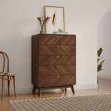 A LuxenHome Theo mid-century modern walnut finish 5-drawer chest stands on a hardwood floor. A framed feather artwork and a vase with flowers are on top. A wooden chair is on the left, a plant is partially visible on the right, and a patterned rug lies in front.