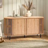 A wooden mid-century modern buffet cabinet with a slatted design stands on black legs. It features an oak wood grain finish and is adorned with vases and a book, set on a light wood floor against panelled walls.