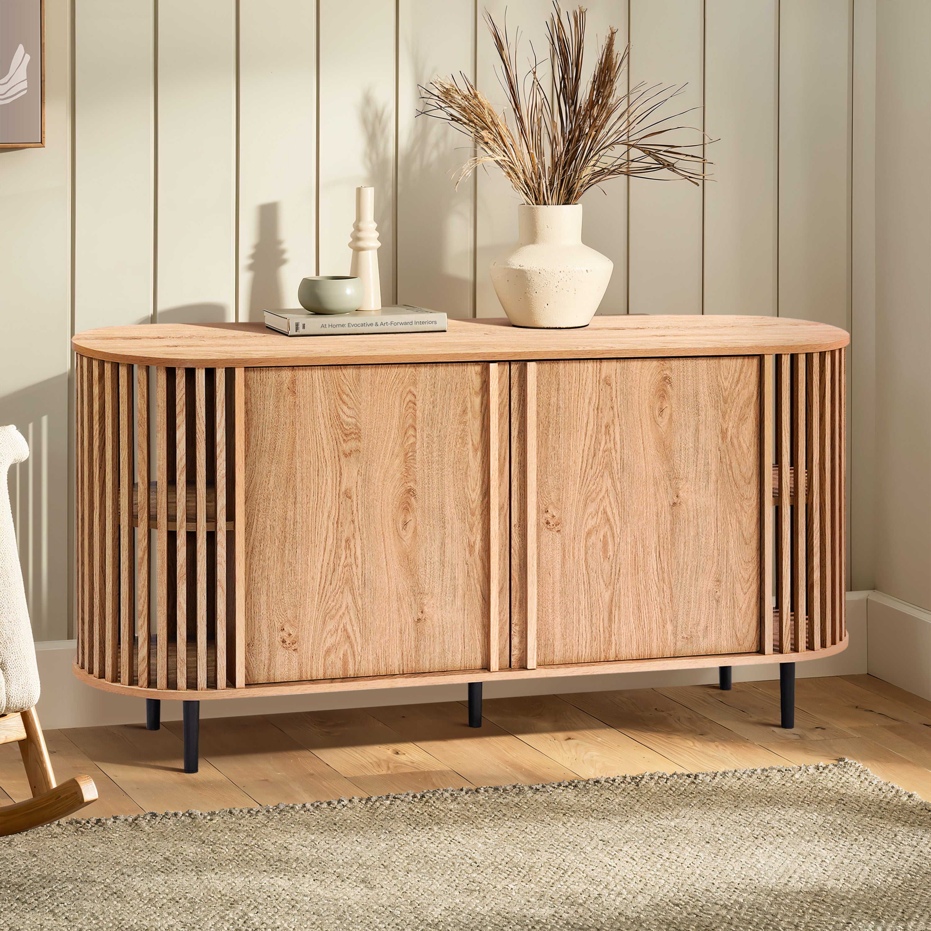 A wooden mid-century modern buffet cabinet with a slatted design stands on black legs. It features an oak wood grain finish and is adorned with vases and a book, set on a light wood floor against panelled walls.
