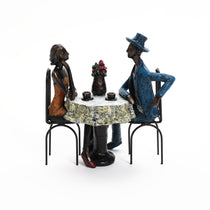 Two abstract figurines, a man in a blue outfit and a woman in an orange dress, capture romantic moments at a round table adorned with floral centerpiece and coffee cups—a modern brown resin couple decoration perfect as a wedding gift.