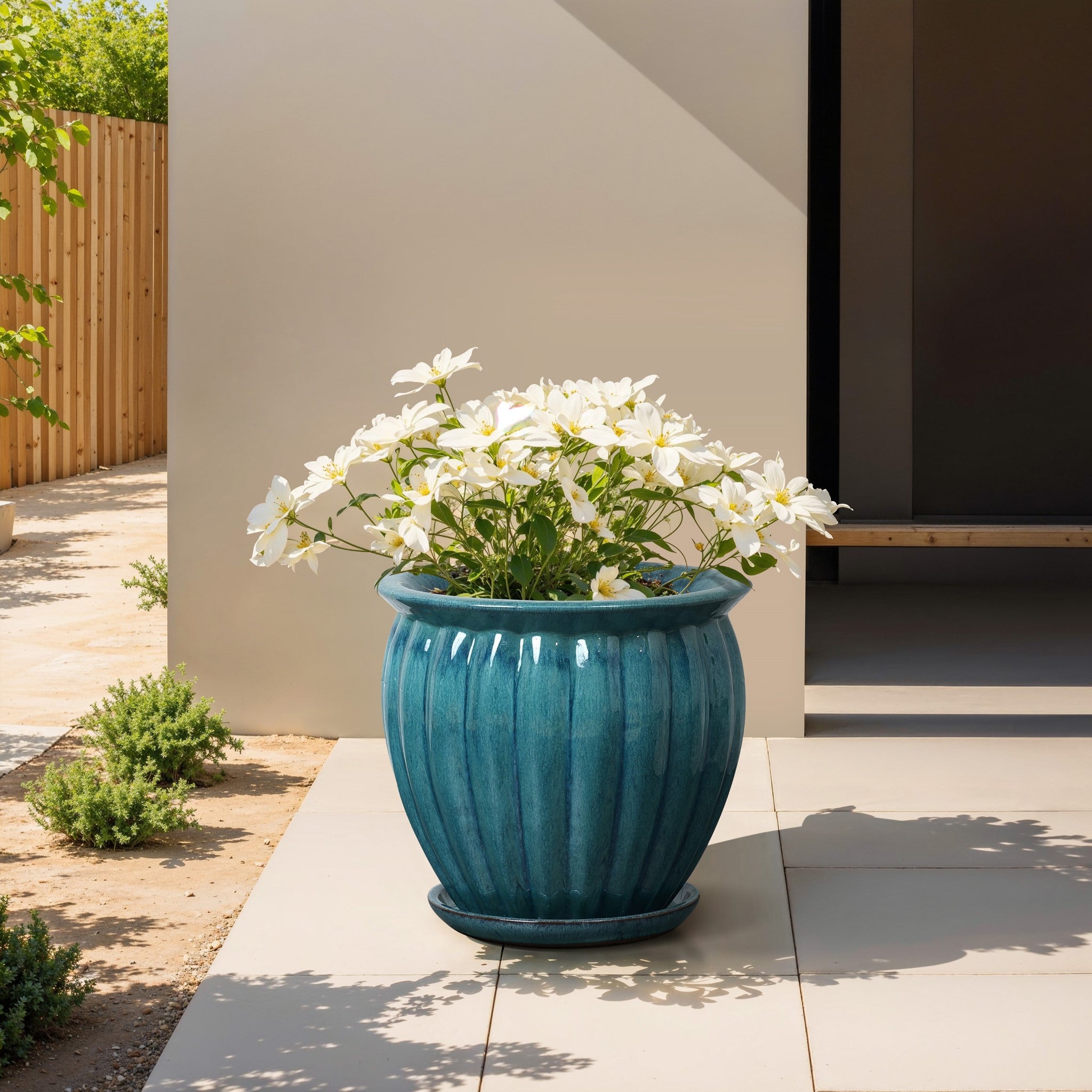 A ceramic decorative planter with a navy blue glaze and white flowers sits on a sunny patio, surrounded by a wooden fence and green shrubs nearby.
