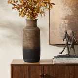 A LuxenHome brown ombre terracotta tall vase, 14.8 h, adorns a wooden cabinet with autumnal leaves. Beside it are books and a small sculpture of two figures walking, perfectly complementing the large abstract painting on the wall behind.