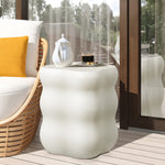 A curvy side table in white, adorned with a coffee mug and pitcher, sits beside a wicker chair with a yellow pillow. Positioned on a wooden deck with glass doors, this scene offers serene views of trees outside—an epitome of modern home furnishings.