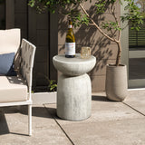 A modern patio setup showcases a sleek white chair and LuxenHomes rustic off-white and gray outdoor side table with a wine bottle and glass. Two tall potted plants complement the scene, set against a taupe wall and concrete floor for an elegant charm.